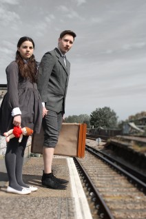A boy and a girl stand on a railway platform, clutching a doll and suitcase. They are looking down the tracks with worried expressions.